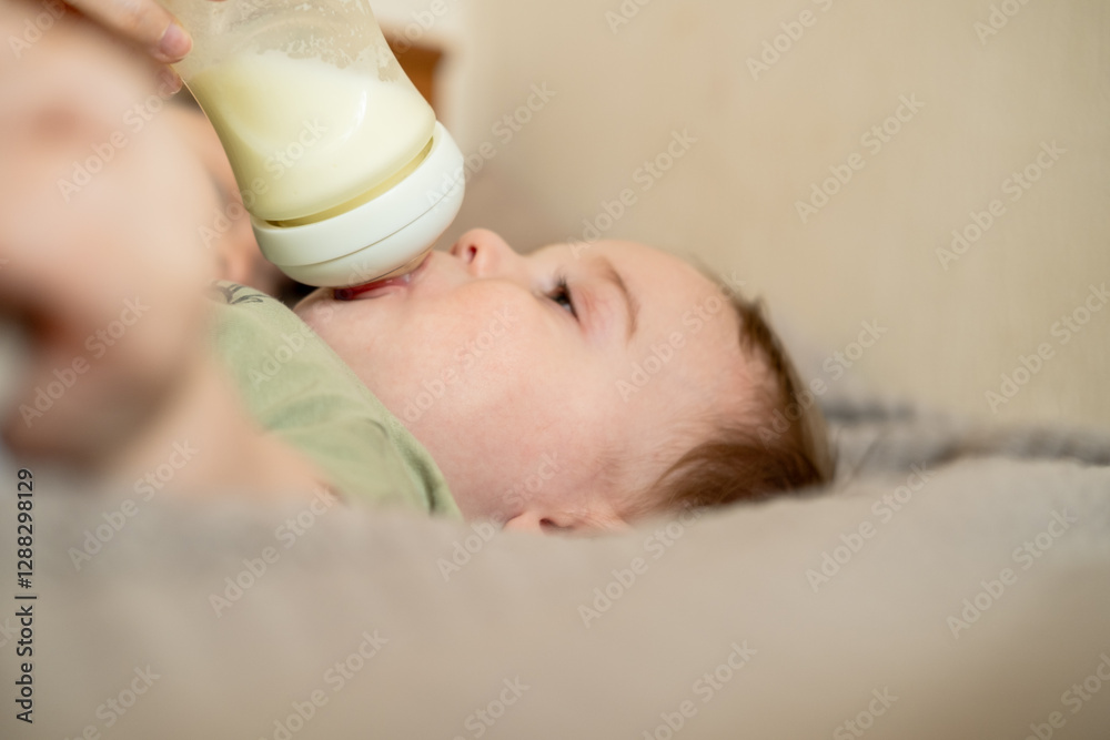 Happy baby boy is drinking milk formula from a bottle lying on the bed. Kid aged 1 year old Happy baby boy is drinking milk formula from a bottle lying on the bed. Kid aged 1 year old