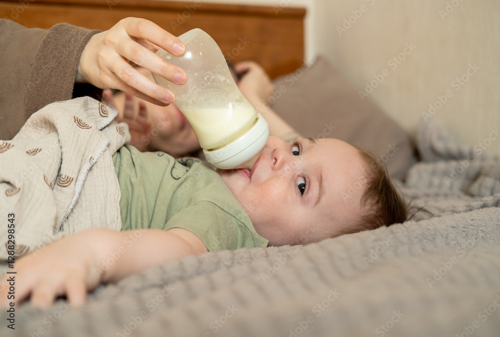Happy baby boy is drinking milk formula from a bottle lying on the bed. Kid aged 1 year old Happy baby boy is drinking milk formula from a bottle lying on the bed. Kid aged 1 year old