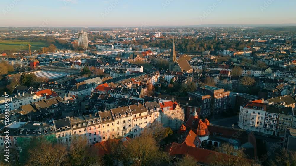 A scenic aerial view of Aachen, featuring red-roofed historic buildings, modern city expansion, green spaces, and distant industrial areas under a clear sky. Aachen, Deutschland A scenic aerial view of Aachen, featuring red-roofed historic buildings, modern city expansion, green spaces, and distant industrial areas under a clear sky. Aachen, Deutschland