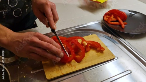 Close-up of a left-handed man slicing red bell peppers on a wooden cutting plate in his home kitchen