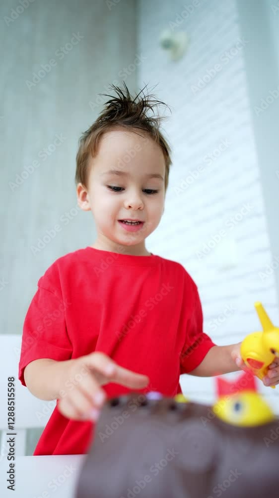Adorable funny Caucasian kid with messy hair in red t-shirt plays at the desk. Low angle view at the toddler involved in a game with toys. Vertical video. Adorable funny Caucasian kid with messy hair in red t-shirt plays at the desk. Low angle view at the toddler involved in a game with toys. Vertical video.