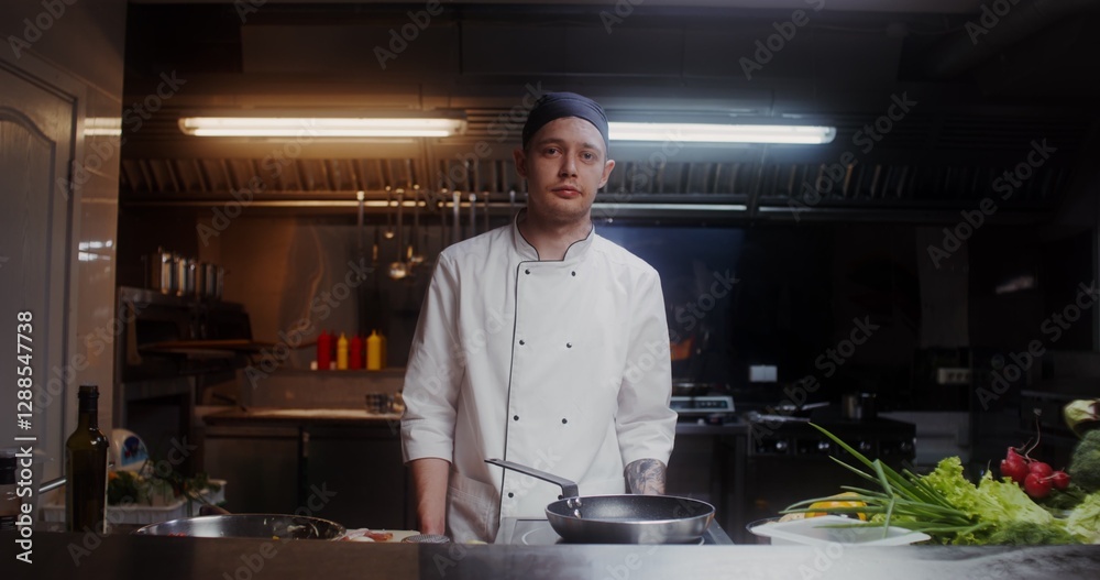 A man in a chef's uniform smiles while looking at the camera standing in the restaurant kitchen A man in a chef's uniform smiles while looking at the camera standing in the restaurant kitchen