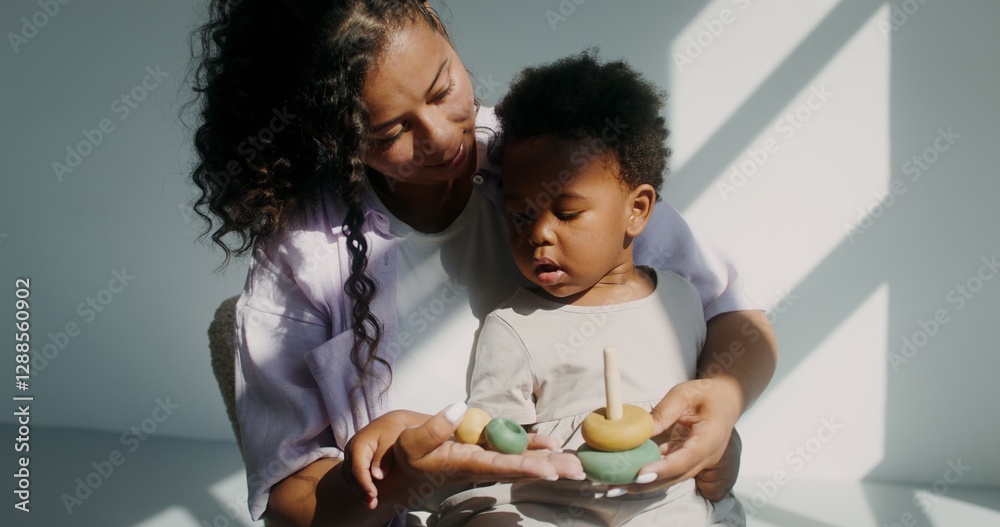 Mom with a child of African-American appearance, dressed in white clothes, playing pyramid while sitting in a chair in a white room Mom with a child of African-American appearance, dressed in white clothes, playing pyramid while sitting in a chair in a white room