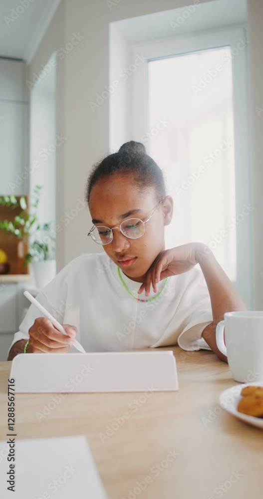 A girl with glasses enthusiastically draws on a digital tablet using a stylus, sitting at a table at home A girl with glasses enthusiastically draws on a digital tablet using a stylus, sitting at a table at home