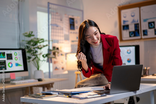 A woman in a red jacket is sitting at a desk with a laptop and smiling