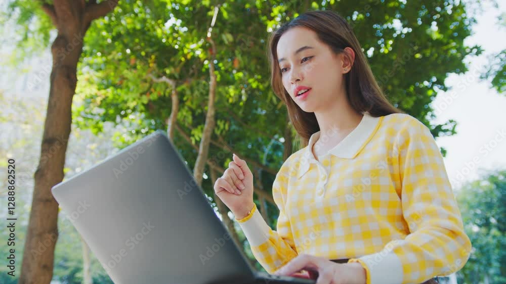 Young woman in yellow checkered shirt working on laptop outdoors, surrounded by nature, expressing excitement and embracing technology in casual setting. Digital nomad remote work