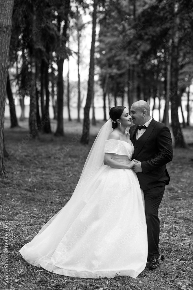 Newlyweds in the forest. The groom is in a black classic suit with a bow tie, and the bride is in a white wedding dress with a long veil. They hold hands and look at each other. Newlyweds in the forest. The groom is in a black classic suit with a bow tie, and the bride is in a white wedding dress with a long veil. They hold hands and look at each other.