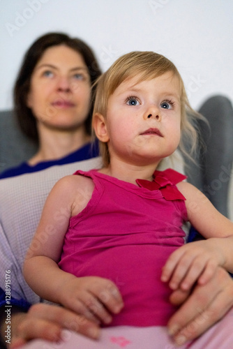 Portrait of a littie girl watching TV while sitting on her mother's lap in the living room of the house. Mom and daughter watching TV.