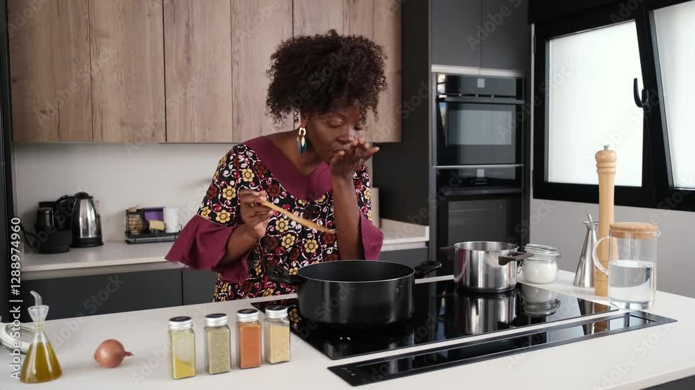 African woman cooks okra soup with spinach, trying it, adding salt and stirring in a pot on a modern kitchen stove