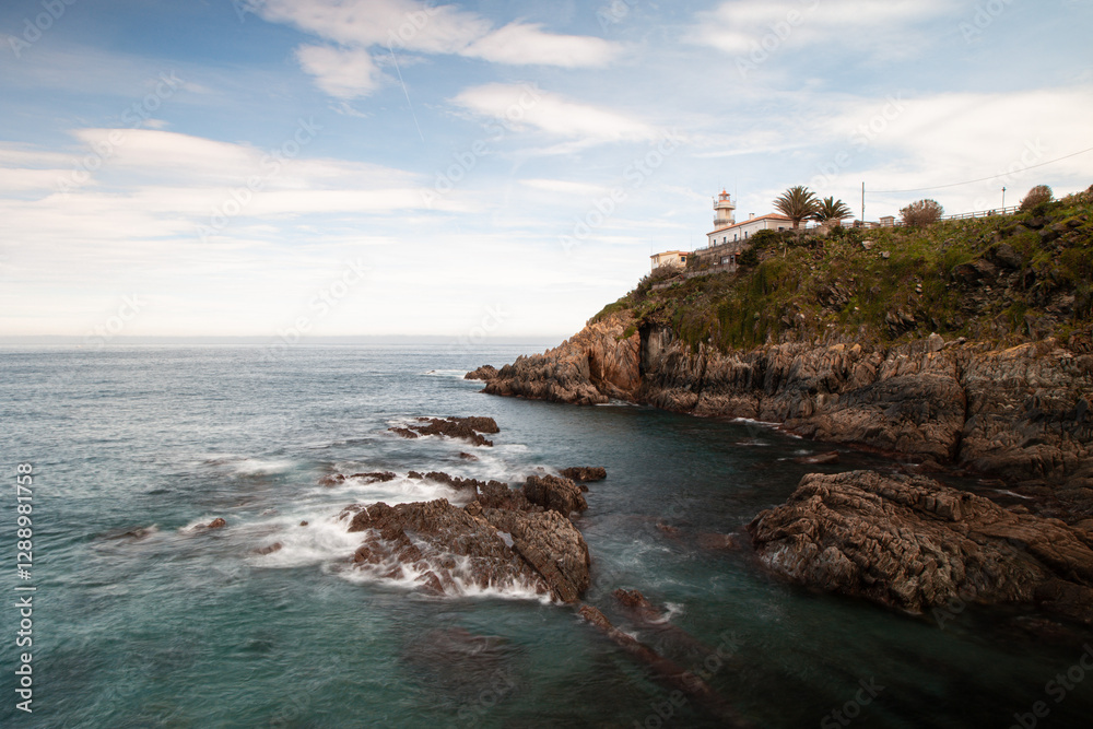 Scenic coastal view of a lighthouse on a rocky cliff overlooking the Atlantic Ocean in Cudillero, Asturias, Spain, with waves crashing against jagged rocks under a partly cloudy sky Scenic coastal view of a lighthouse on a rocky cliff overlooking the Atlantic Ocean in Cudillero, Asturias, Spain, with waves crashing against jagged rocks under a partly cloudy sky