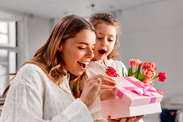 Happy Mother's Day! A happy mother and her excited daughter open a pink gift box together. The mother holds a bouquet of  tulips while both smile with delight in a bright, cozy home setting.