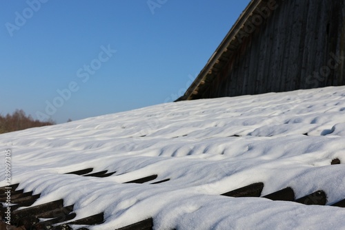 A rustic barn roof covered in a thick layer of undisturbed snow under a clear blue sky. A peaceful winter rural scene