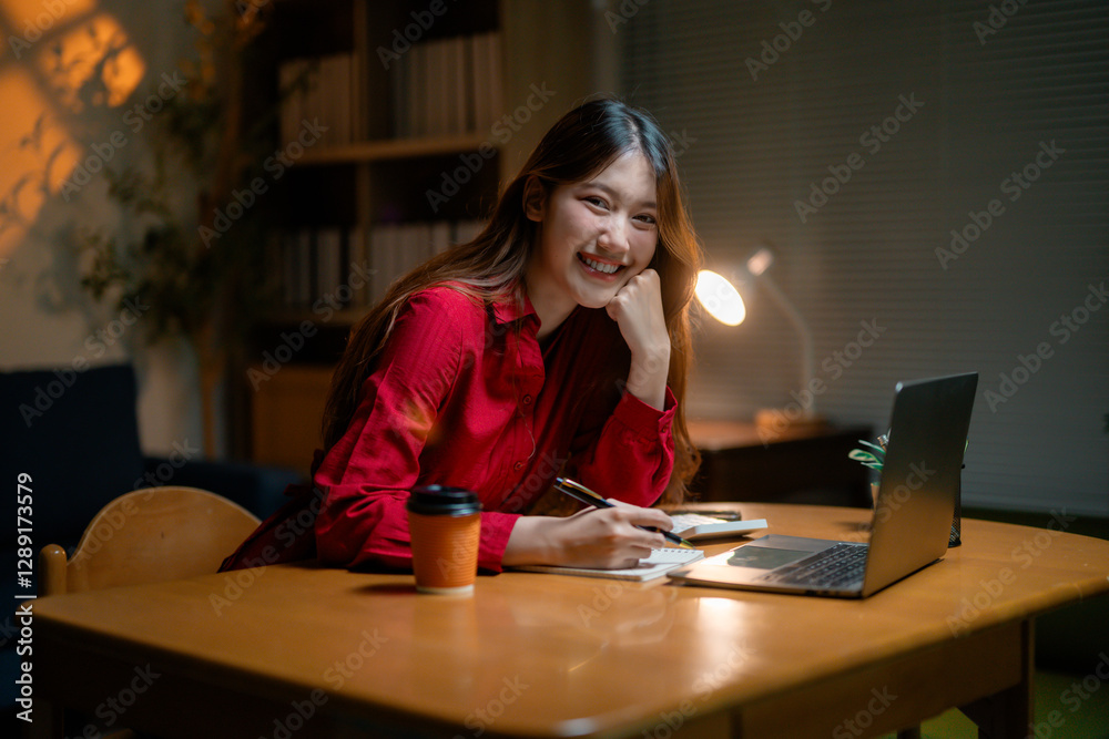 Asian businesswoman working late, typing on laptop and writing notes. Takeaway coffee nearby. Displaying confident, focused expression while reviewing professional documents Asian businesswoman working late, typing on laptop and writing notes. Takeaway coffee nearby. Displaying confident, focused expression while reviewing professional documents