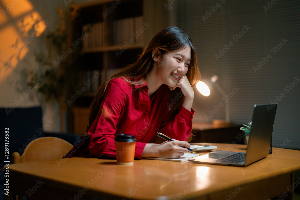 Professional Asian businesswoman wearing red shirt writing notes during late-night video conference, working remotely from home office with laptop Professional Asian businesswoman wearing red shirt writing notes during late-night video conference, working remotely from home office with laptop