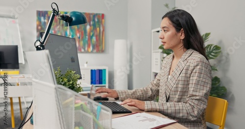 An attractive, experienced company accountant sits in office in front of a computer checking statistical data employee salary charts comparing paper diagrams with electronic ones financial analyst