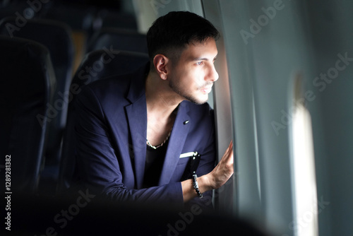 Handsome business man sitting near window in cabin during flight looking out through airplane window.