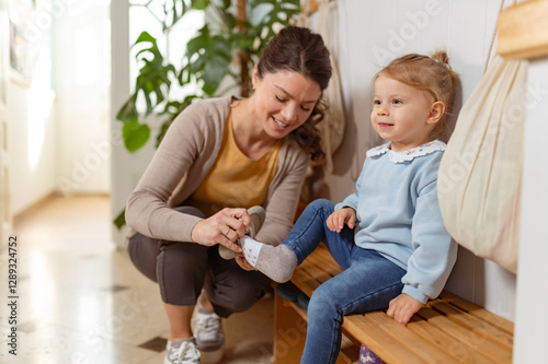 A young woman helps a little girl put on her shoes in a preschool or daycare dressing area, showing care and support in a warm, nurturing environment
