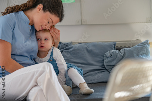A caregiver in a blue uniform gently kisses a sick young girl's forehead while comforting her on a couch, showing warmth and care in a classroom or daycare setting