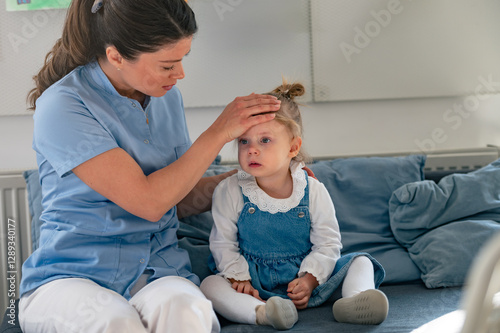 A concerned caregiver checks a young girl's forehead for fever as she sits on a couch in a classroom, looking unwell and in need of care and comfort