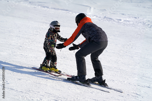 Teaching a child to ski on alpine skis. A man backs up the baby on a snowy slope.