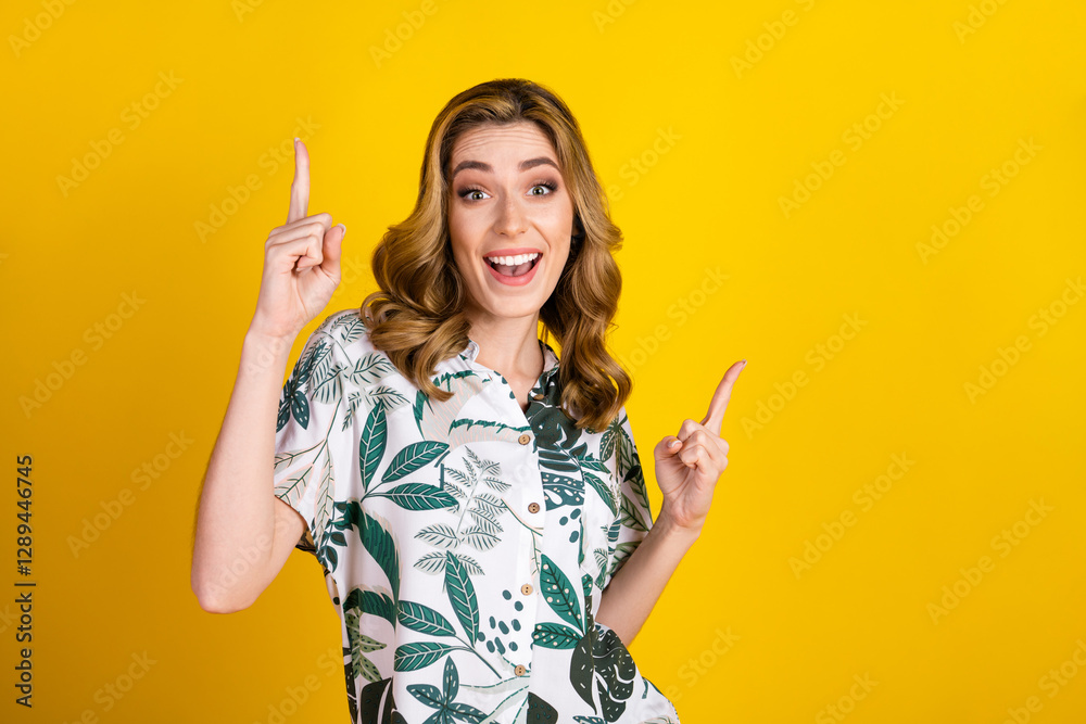 Smiling woman in floral shirt pointing upwards on vibrant yellow background expressing joy and making cheerful hand gestures Smiling woman in floral shirt pointing upwards on vibrant yellow background expressing joy and making cheerful hand gestures