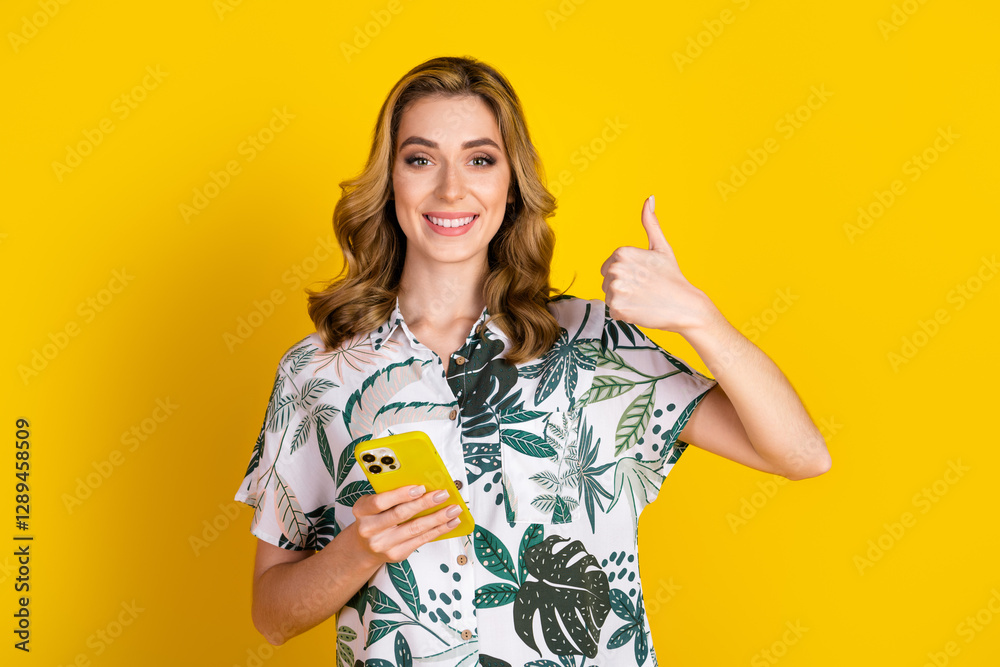Smiling woman in floral shirt holding phone with thumbs-up gesture against bright yellow background Smiling woman in floral shirt holding phone with thumbs-up gesture against bright yellow background