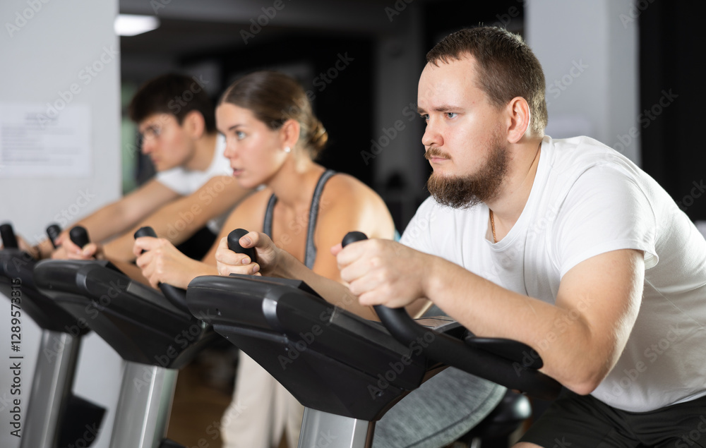 Bearded man hardly perform exercise on stationary bicycle, pedaling and exercising in fitness class. Fitness equipment, visitors engaged in physical task during training Bearded man hardly perform exercise on stationary bicycle, pedaling and exercising in fitness class. Fitness equipment, visitors engaged in physical task during training