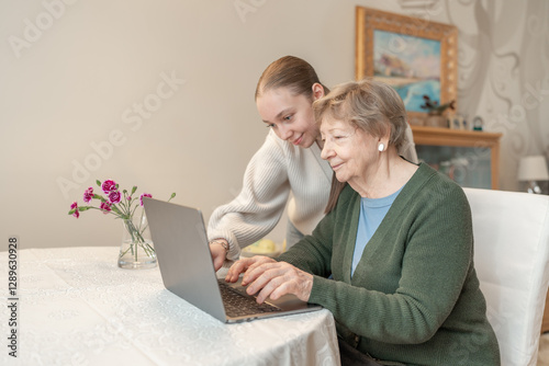 Young woman helping a senior woman with using laptop at home. Concept of technology, education, and senior digital adaptation. Granddaughter teaches grandmother how to use digital device.