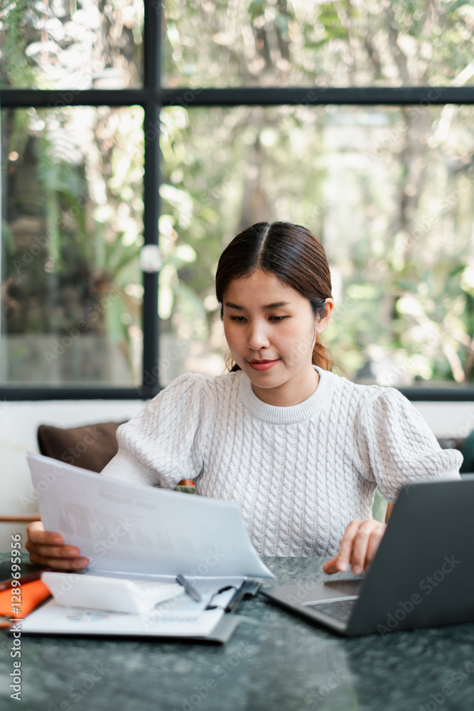 A focused young woman works on her laptop while reviewing documents in a modern, bright living room with large windows and lush greenery outside. A focused young woman works on her laptop while reviewing documents in a modern, bright living room with large windows and lush greenery outside.