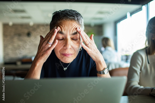 Stressed senior businesswoman massaging temples while working on laptop in office photo