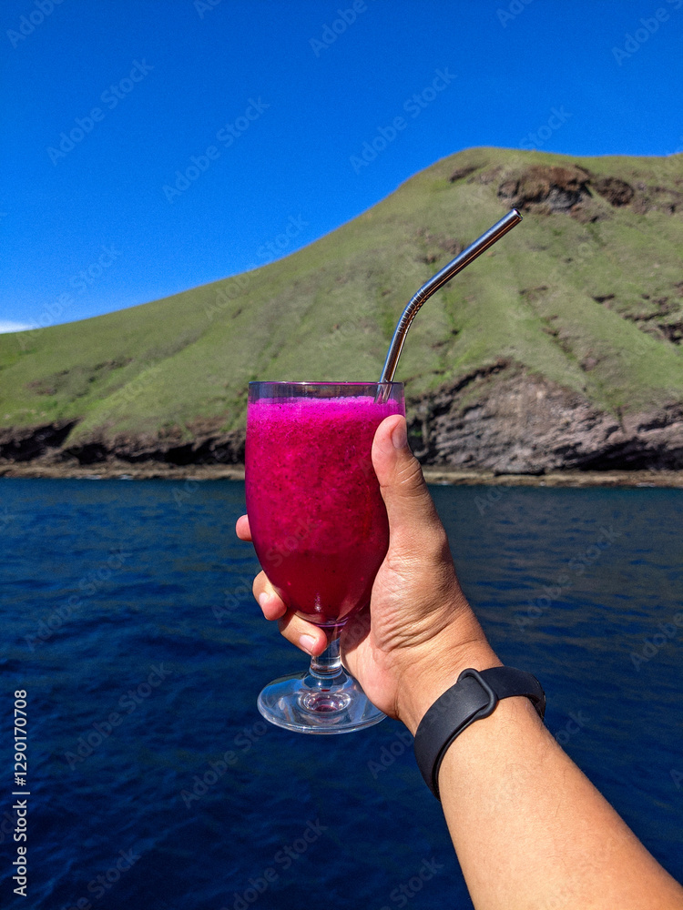 A hand holding a glass of vibrant dragon fruit juice with a metal straw, set against a scenic backdrop of a lush green hill and deep blue ocean under a clear sky.