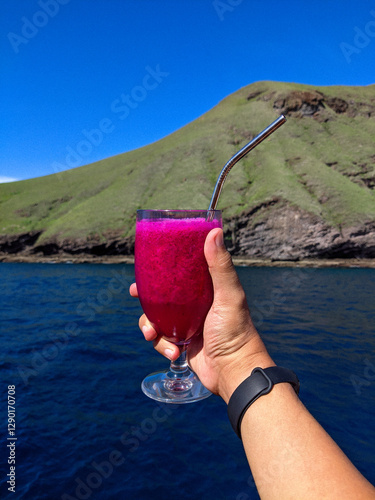 Obraz na płótnie A hand holding a glass of vibrant dragon fruit juice with a metal straw, set against a scenic backdrop of a lush green hill and deep blue ocean under a clear sky. w salonie