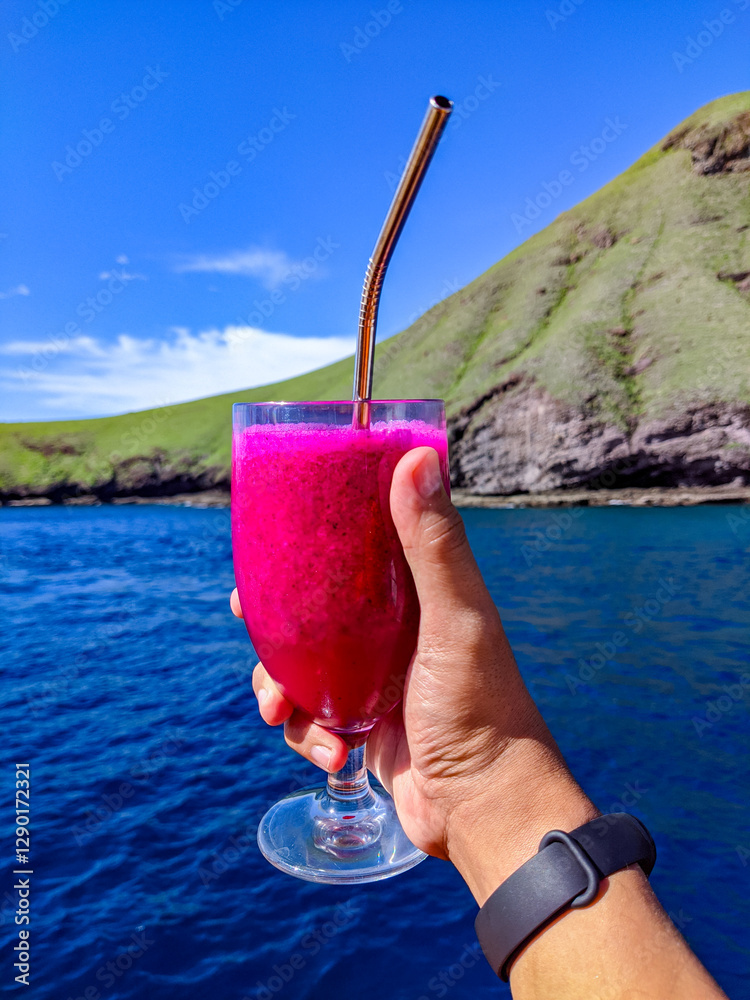 A hand holding a glass of vibrant dragon fruit juice with a metal straw, set against a scenic backdrop of a lush green hill and deep blue ocean under a clear sky