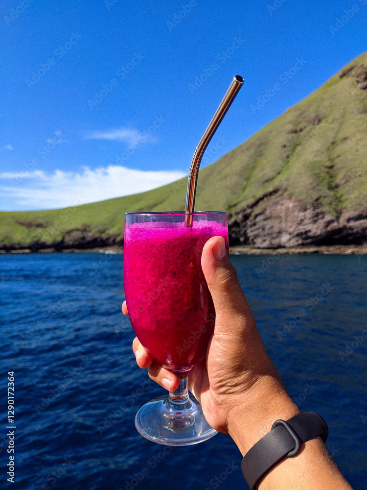 A hand holding a glass of vibrant dragon fruit juice with a metal straw, set against a scenic backdrop of a lush green hill and deep blue ocean under a clear sky