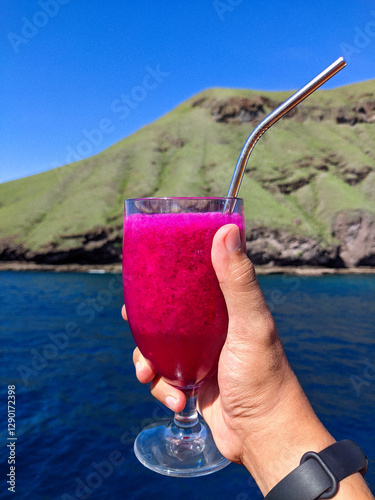 A hand holding a glass of vibrant dragon fruit juice with a metal straw, set against a scenic backdrop of a lush green hill and deep blue ocean under a clear sky