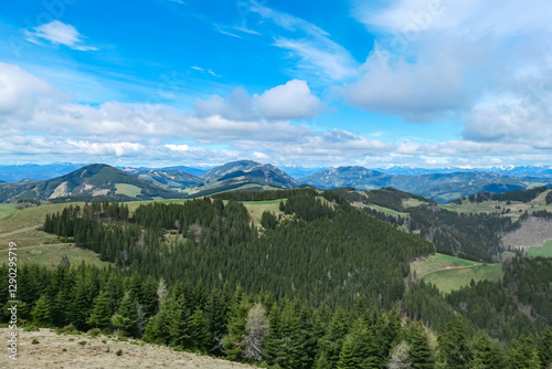 Verdant rolling hills across Plankogel, Styria, Austria, under partly cloudy sky. Patches of coniferous trees dot the landscape, creating a textured, scenic view of the Austrian countryside. Tranquil