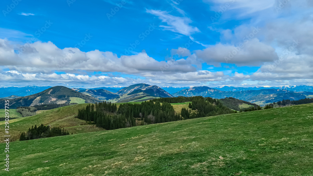 Fototapeta premium Verdant rolling hills across Plankogel, Styria, Austria, under partly cloudy sky. Patches of coniferous trees dot the landscape, creating a textured, scenic view of the Austrian countryside. Tranquil
