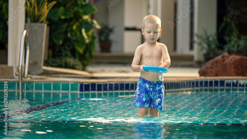 A young child stands in shallow water, holding a blue frisbee while wearing patterned swim shorts. Bright sunlight illuminates the cheerful atmosphere around the pool.