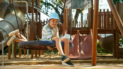 A young boy with a blue hat and striped shirt sits on a tire swing, looking thoughtful. He enjoys a moment of quiet while surrounded by playground equipment on a sunny day.