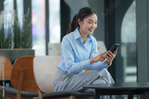 Young asian businesswoman sitting on a chair in a modern office, using her smartphone with a smile, showcasing the concept of mobile technology in the workplace