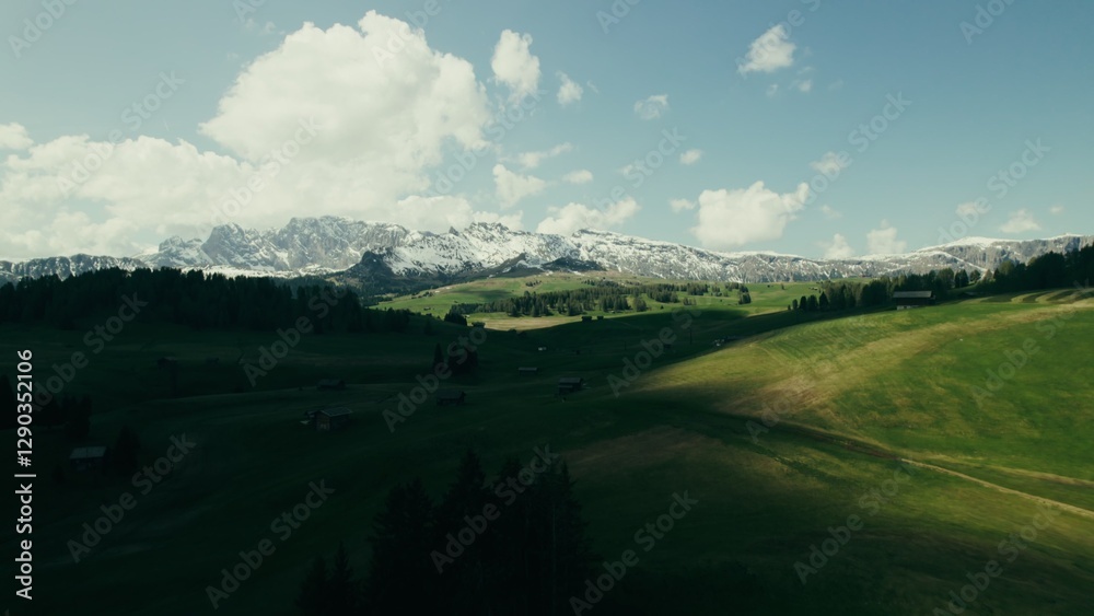 Obraz premium The drone flies over green meadows and rare houses on the hills at the foot of the rocky mountains.