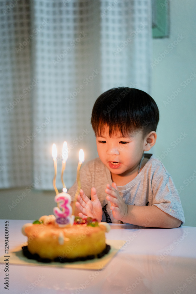 young boy eagerly looks at birthday cake with lit candles, celebrating his third birthday. room is softly lit, creating warm and joyful atmosphere