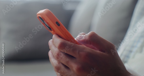 Close-up of a mobile phone with three cameras, in an orange case in men's hands indoors. Man typing on smartphone, no face