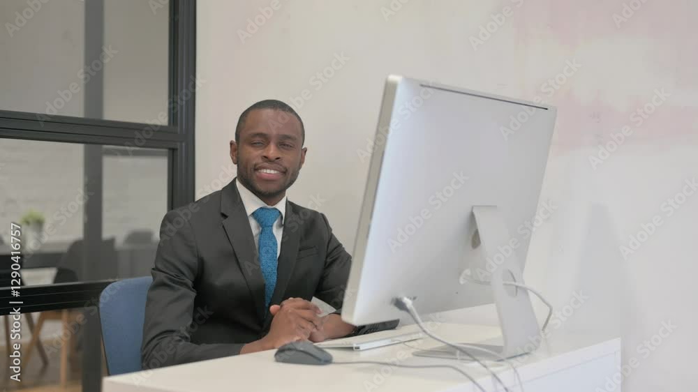 African Businessman Smiling at Camera while Working on Desktop Computer African Businessman Smiling at Camera while Working on Desktop Computer