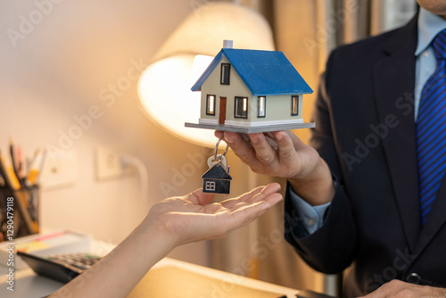 A man is holding a key in his hand and is about to give it to a woman. The woman is holding a small house in her hand. The scene seems to be about a house sale or a housewarming party
