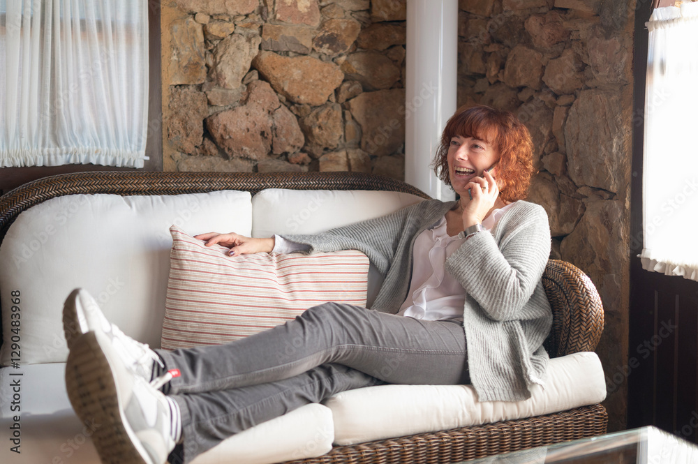 Obraz premium Happy woman relaxing on sofa, talking on mobile phone in rustic living room