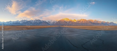 Panoramic view of a sunrise over a mountainous landscape with golden peaks and a clear blue sky above an empty asphalt road in the foreground
