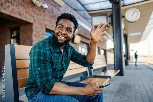 Young black passenger sitting on train platform on a bench with phone in hands and waving to friend.