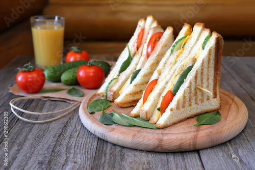 On a wooden table against the log wall chicken sandwiches with greens are on a tray next to a glass of orange juice and fresh vegetables on a paper bag.