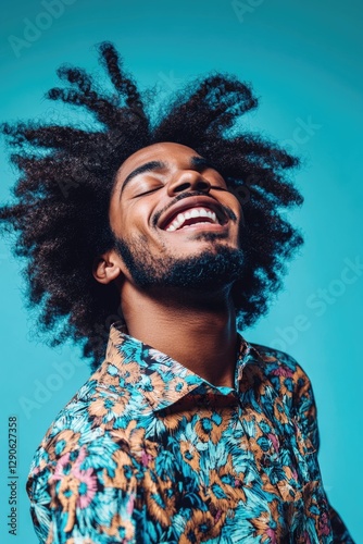 A young man with a beard and dreadlocks, wearing a colorful shirt, laughing heartily against a blue background.